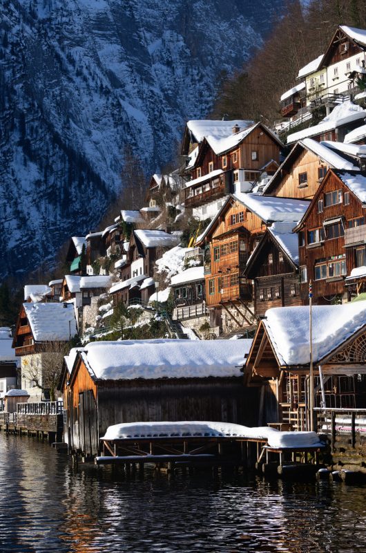 The traditional wooden lakeside houses of Hallstatt town with snow on the roofs in the first sunlight of the morning and with Alps mountains in the background in winter in Gmunden, Upper Austria.